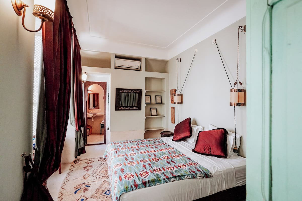 A bedroom with a bed, a wardrobe, a shelf with decorative items, and a door leading to another room.