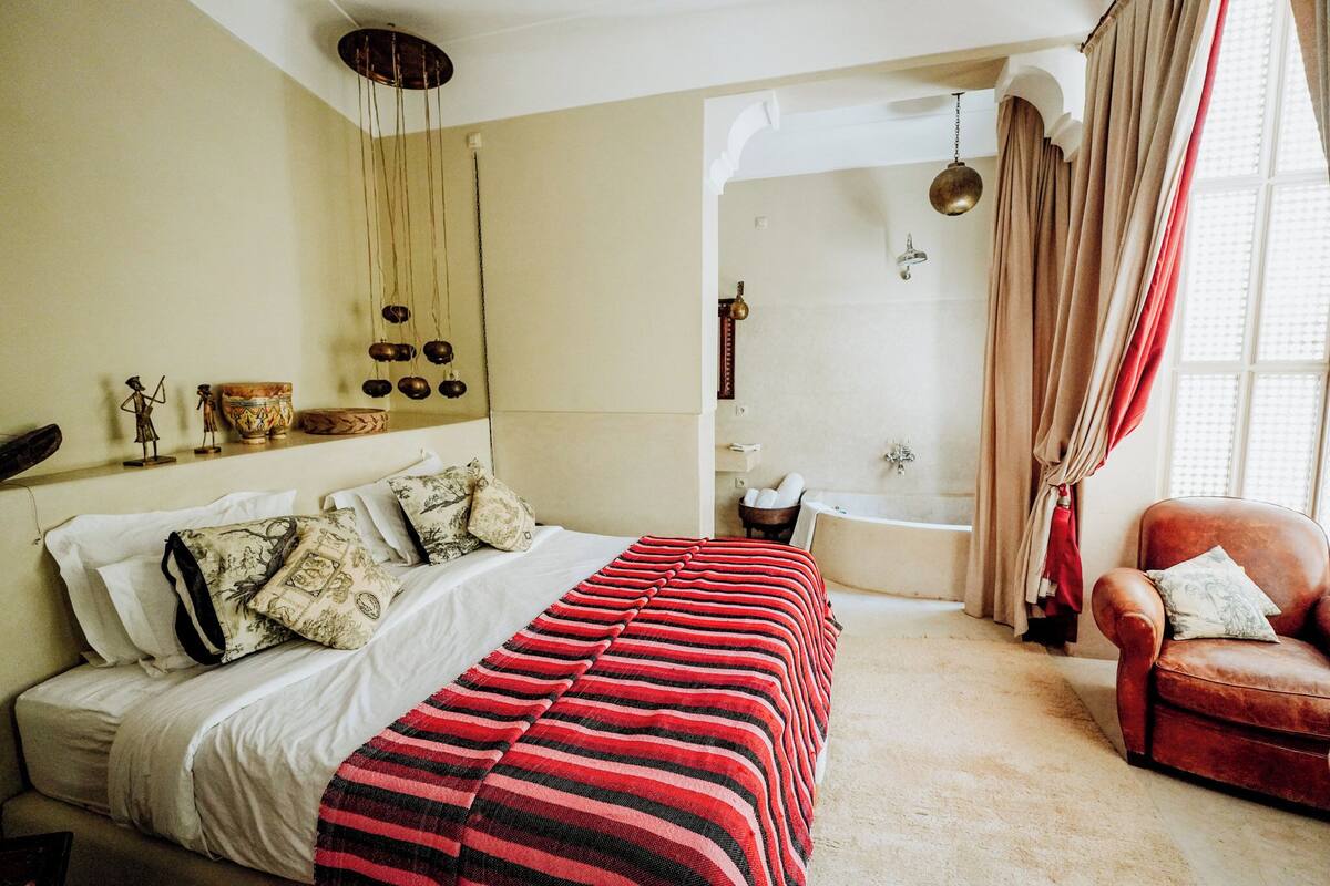 A bedroom with a bed, a red and black striped blanket, a red armchair, a bathtub, and a shower.