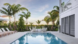 Una piscina con sillones de descanso y una mesa, rodeada de palmeras y una valla.