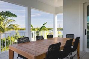 A covered outdoor dining area with a wooden table and black chairs, overlooking a garden with palm trees and a view of the water.