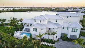 Una casa grande y blanca de varios pisos con piscina, rodeada de palmeras y un puerto deportivo al fondo.