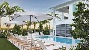 Et poolområde med liggestole og parasoller, en pool samt en toetagers bygning med balkon.