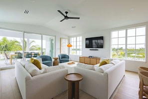 A modern living room with a white sofa set, a coffee table, and a TV mounted on the wall. There is a ceiling fan, large windows, and a balcony with outdoor furniture. - The Joyrise House at Solaire Lunara Bay (Key West)