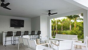 Un espacio moderno para sentarse al aire libre con mobiliario blanco y sillas negras, un televisor de pantalla plana en la pared y vistas a exuberante vegetación.