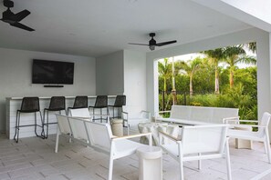 Un espacio moderno para sentarse al aire libre con mobiliario blanco y sillas negras, un televisor de pantalla plana en la pared y vistas a exuberante vegetación.