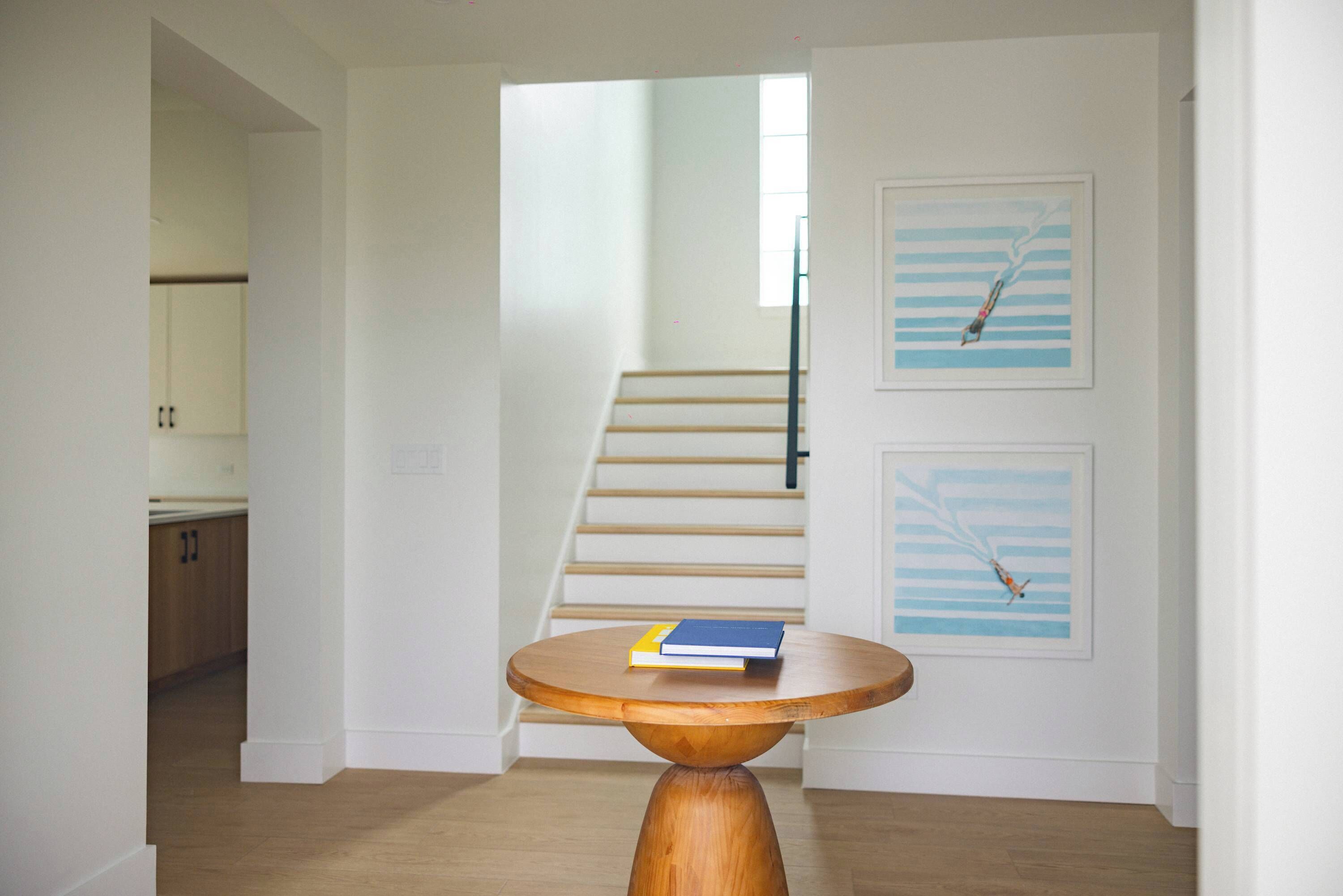 A hallway with a wooden staircase, a round wooden table with books, and two framed artworks on the wall.
