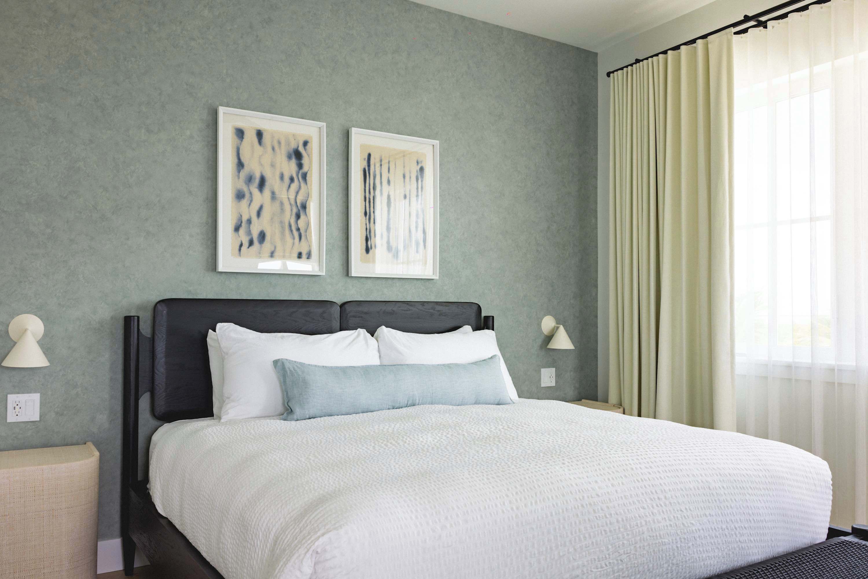 A neatly made bed with white linens, two wall-mounted artworks, and a window with sheer curtains.