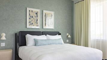 A neatly made bed with white linens, two wall-mounted artworks, and a window with sheer curtains.