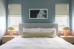 Ein Schlafzimmer mit einem großen Bett, zwei Nachttischlampen, einem gerahmten Bild an der Wand und zwei Fenstern mit Jalousien.
