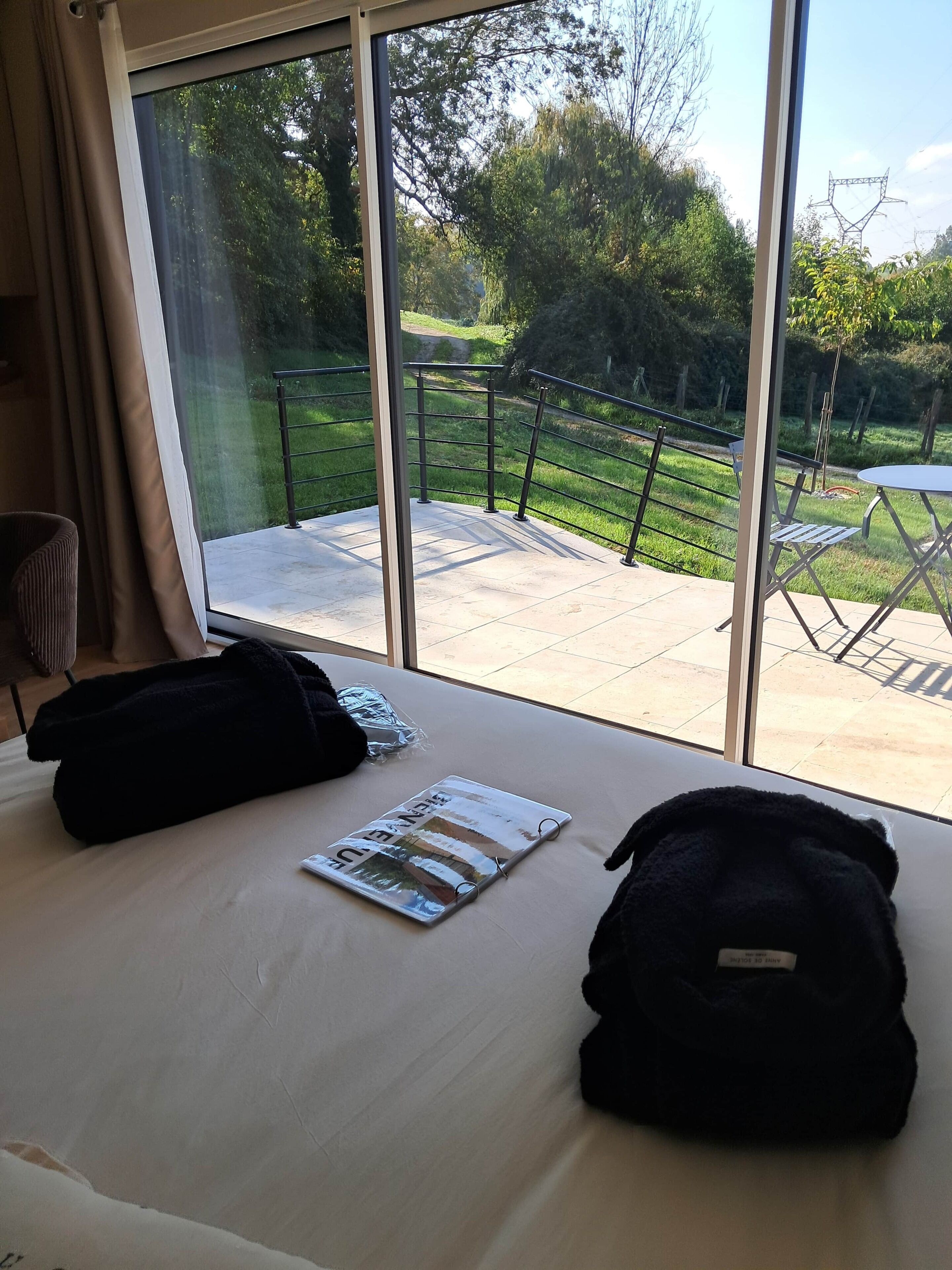 Une chambre d’hôtel avec une grande fenêtre, un lit avec un magazine et un sac noir, et une vue sur un jardin et un espace de détente extérieur.