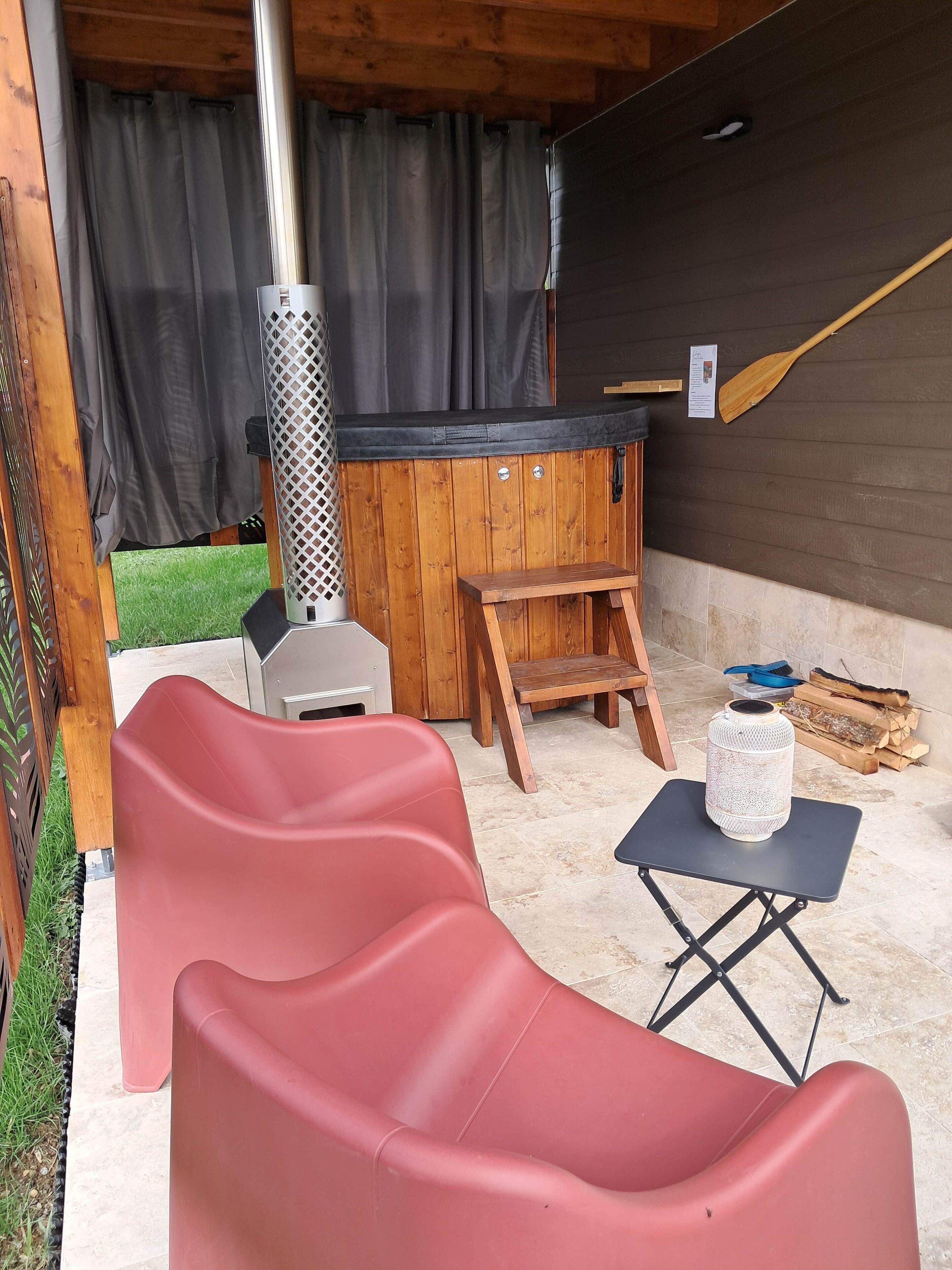 A covered outdoor seating area with a barbecue grill, a wooden table, and a red plastic chair.