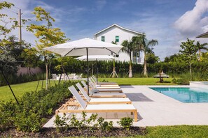 Een zwembadterras met ligbedden en parasols, een zwembad en een wit huis op de achtergrond.
