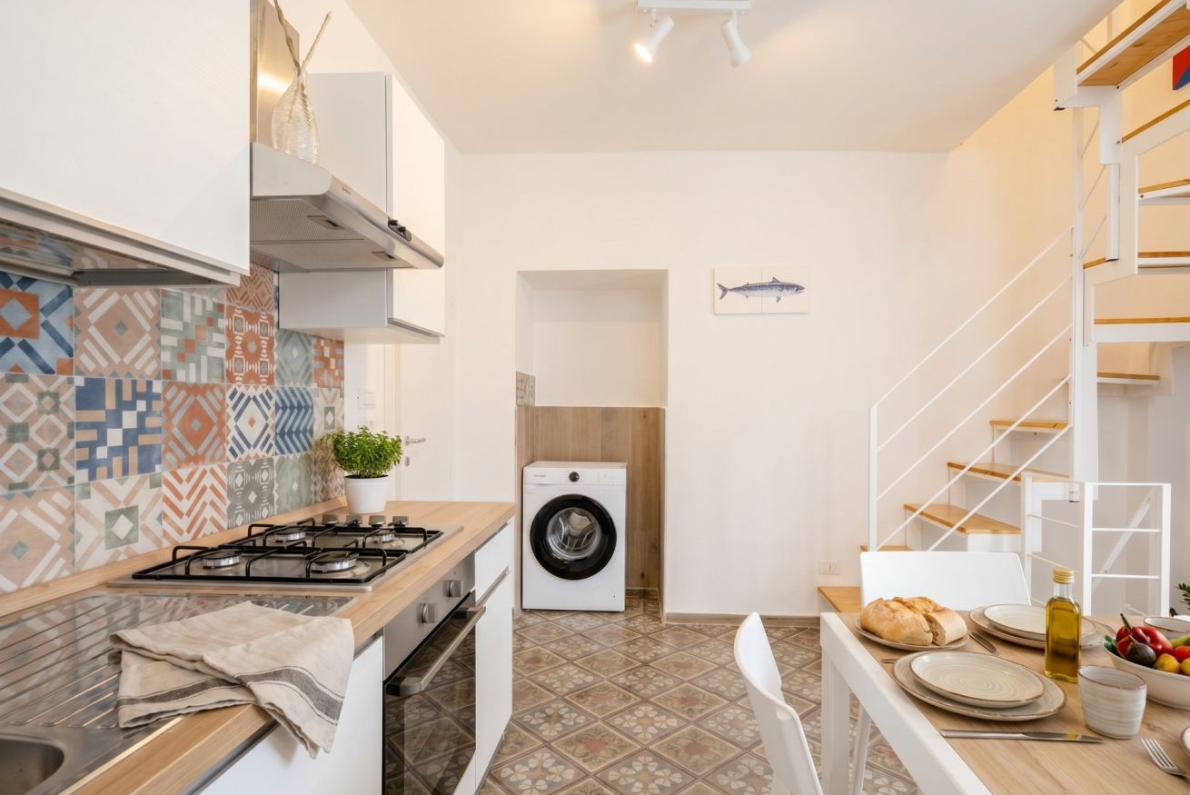 Une cuisine compacte équipée d’un lave-linge, un coin repas avec une table pour deux personnes et un escalier avec une rampe blanche.