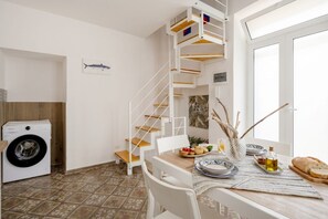 A compact kitchen with a washing machine, a dining table set with plates, a bottle of oil, and a loaf of bread.