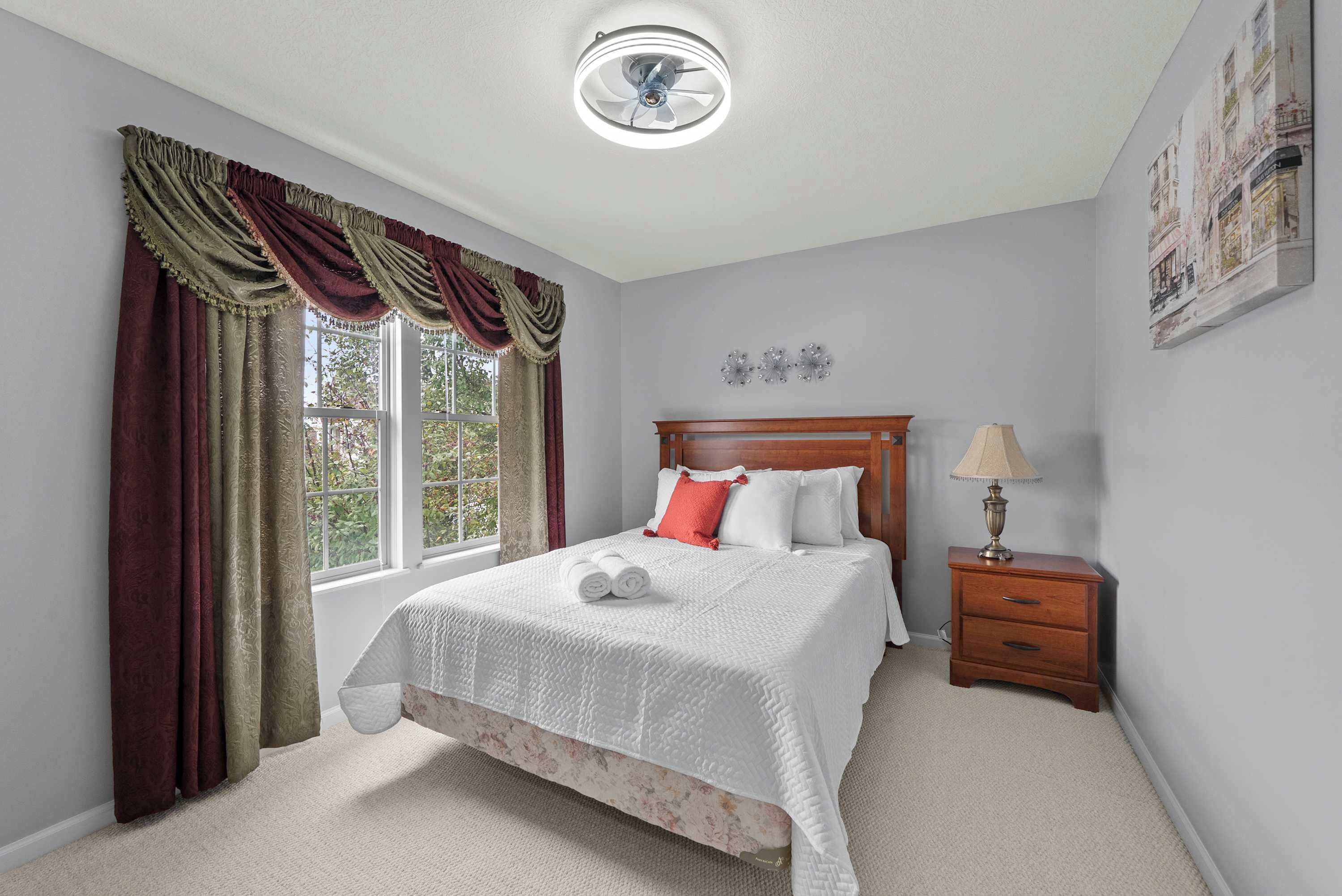 A bedroom with a bed, a nightstand with a lamp, a window with curtains, and a ceiling fan.