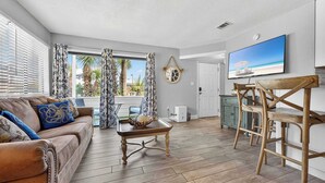 A living room with a sofa, coffee table, and wooden chairs. There is a TV mounted on the wall and a door leading to another room. - Miramar Beach Oasis | Pool & Beach Nearby (Miramar Beach)