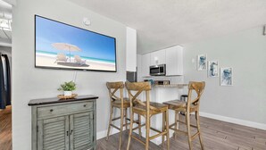 A modern kitchen with white cabinets, a microwave, and a TV displaying a beach scene. There are four wooden chairs around a small dining table. - Miramar Beach Oasis | Pool & Beach Nearby (Miramar Beach)