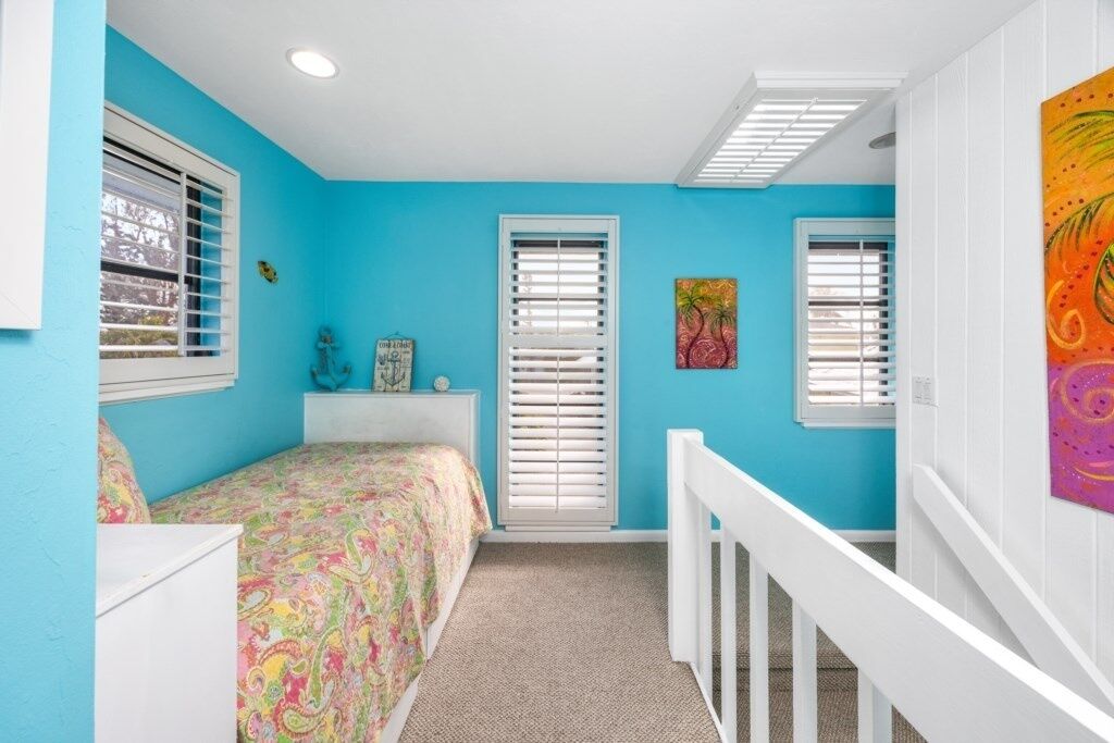 Un dormitorio con cama, dos ventanas con persianas, una cuna blanca y un cuadro colorido en la pared.