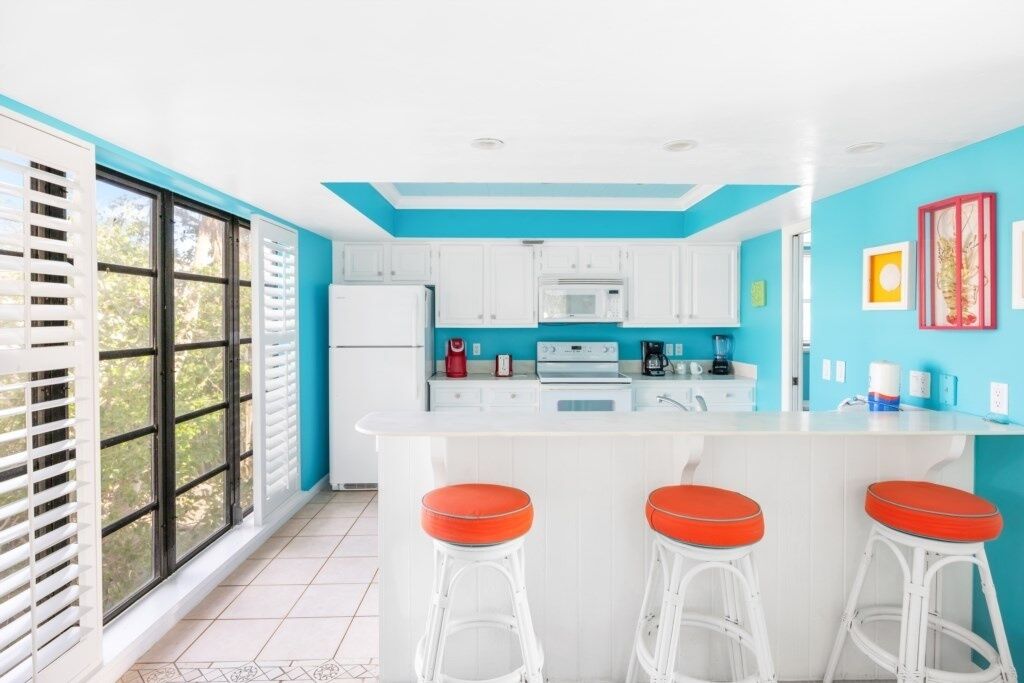 A kitchen with white cabinets, a central island with stools, and a view through large windows.