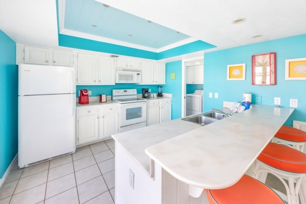 A modern kitchen with white cabinets, a large island with orange stools, a microwave, and a refrigerator.