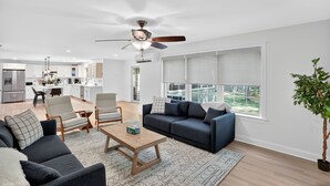 A modern living room with a sofa, coffee table, and chairs, a kitchen with white cabinets, and a large window with blinds. - Gray Woodland Escape Golf, Pool & Cinema 5 Bedroom Home (Gray)