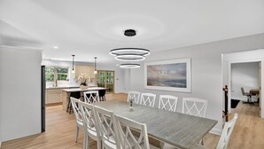 A modern dining area with a long table and white chairs, a kitchen in the background, and a large window. - Gray Woodland Escape Golf, Pool & Cinema 5 Bedroom Home (Gray)