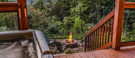 Un bain à remous sur une terrasse en bois, avec un foyer au centre, le tout entouré d’un paysage boisé.