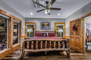 A bedroom with a rustic wooden bed, large windows, a ceiling fan, and a framed picture on the wall.