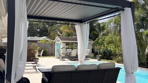 A covered outdoor seating area with a pool, featuring a black wicker sofa and white curtains. - Luxury Pool Dog Friendly Central To Naples And Marco Island By Sky Run Naples 3 Bedroom Home (Naples)