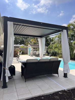 A covered outdoor seating area with a pool, featuring a black wicker sofa and white curtains. - Luxury Pool Dog Friendly Central To Naples And Marco Island By Sky Run Naples 3 Bedroom Home (Naples)
