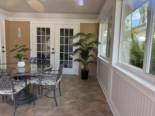 A dining area with a glass table and chairs, a potted plant, and a view of palm trees through a window. - Serendipity At Naples Park By Sky Run Naples 3 Bedroom Home (Naples)