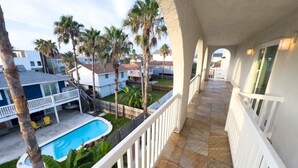 A balcony with a view of a swimming pool, palm trees, and neighboring houses. - Fun Retro Island Vibe Sleeps 17 Beach 1 Block 4 Bedroom Home (South Padre Island)