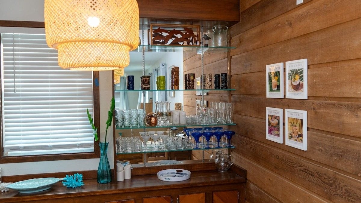 Zona de bar con estantes de vidrio exhibiendo diferentes copas y una lámpara colgante.