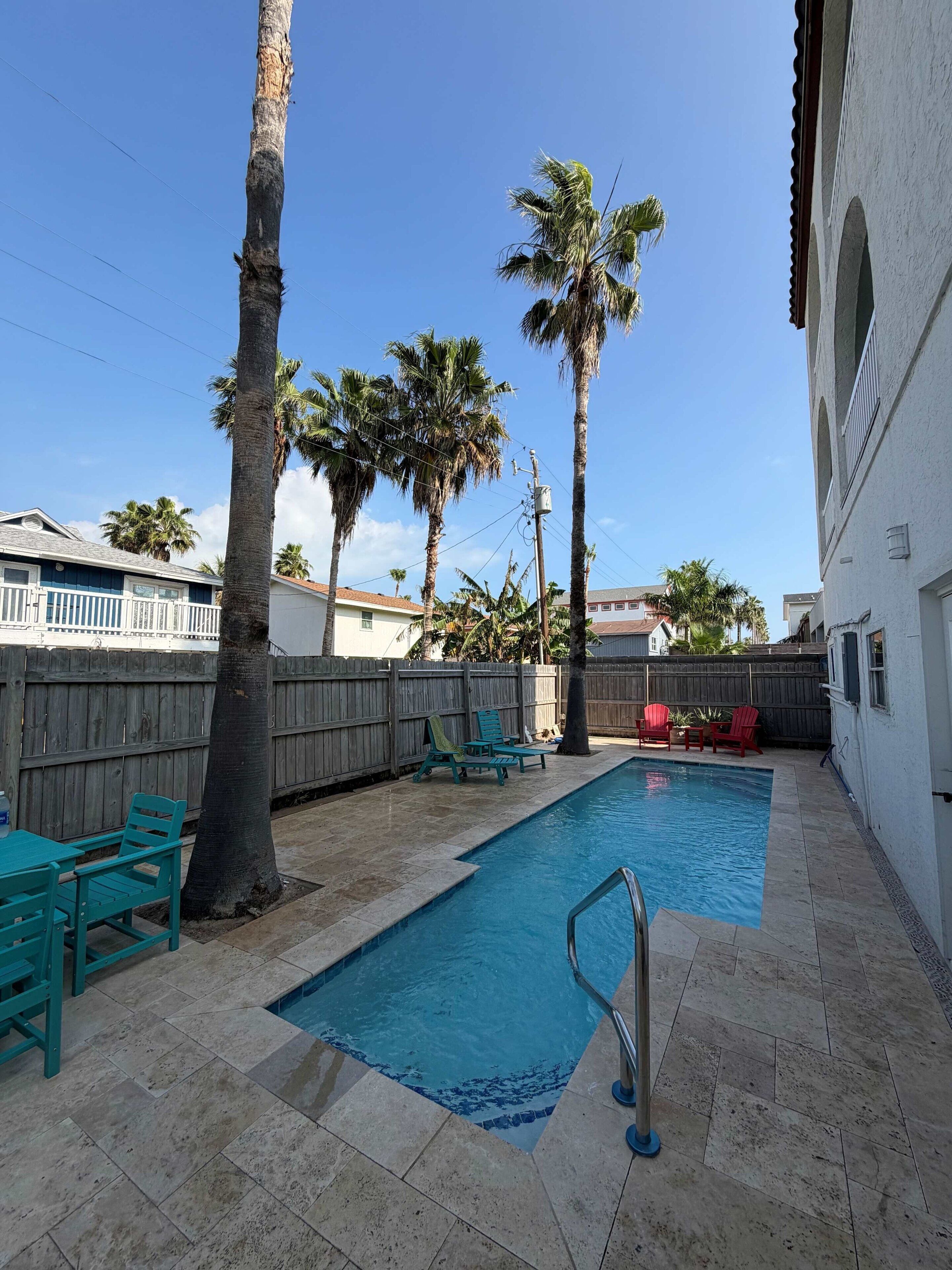 A swimming pool with a tiled deck, surrounded by palm trees and a wooden fence.