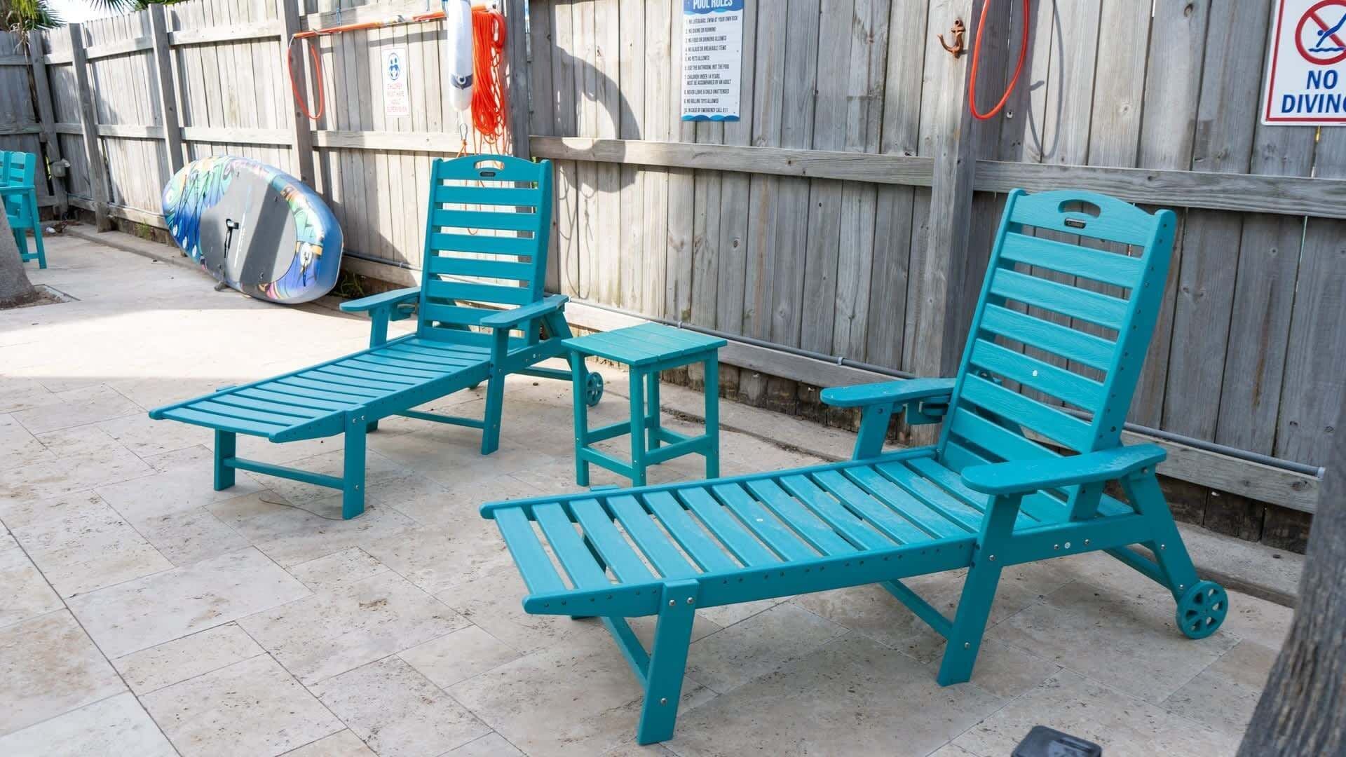 Two turquoise lounge chairs and a small table in a shaded outdoor area with a wooden fence in the background.