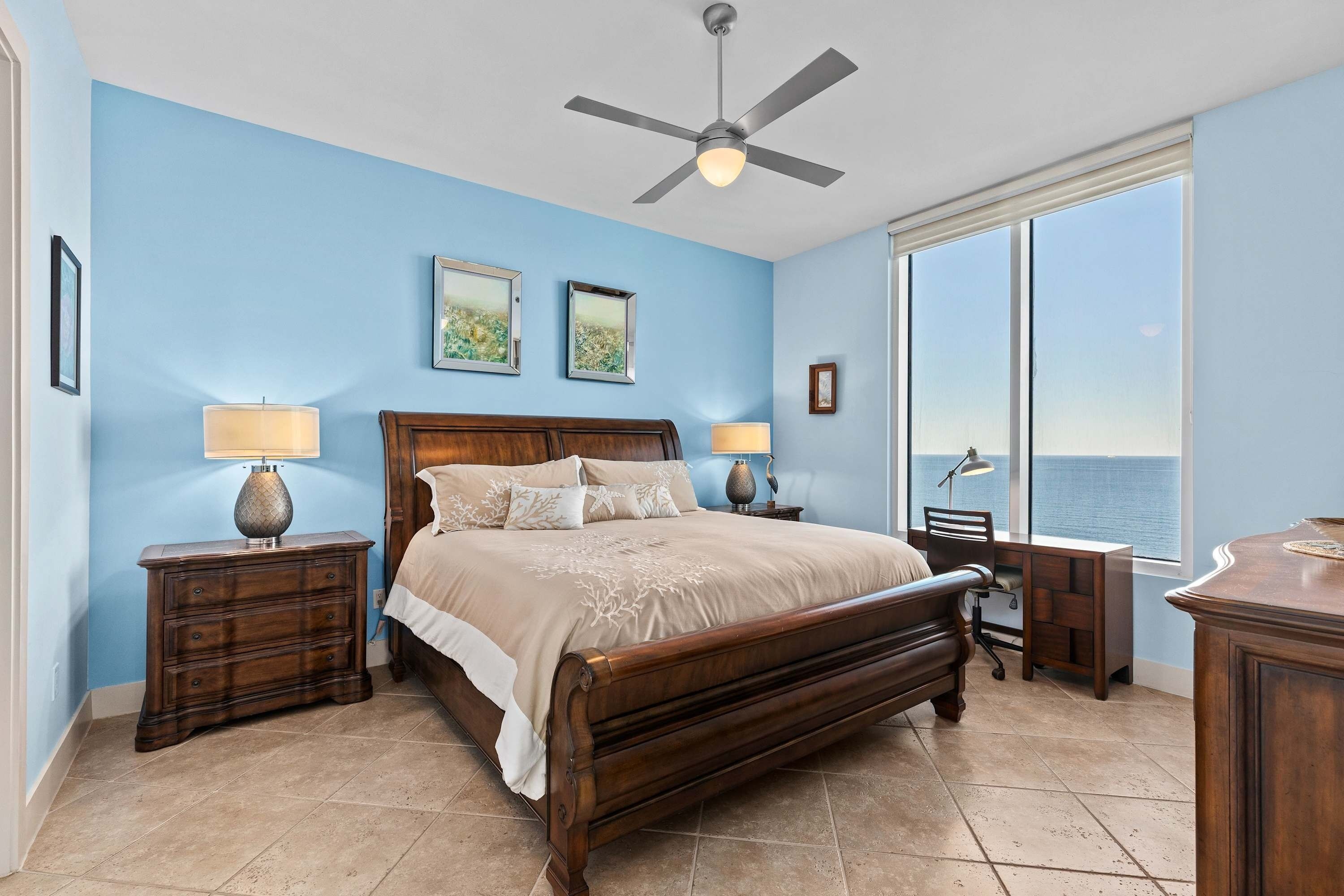 A bedroom with a large bed, two bedside tables, a desk, and a view of the ocean.