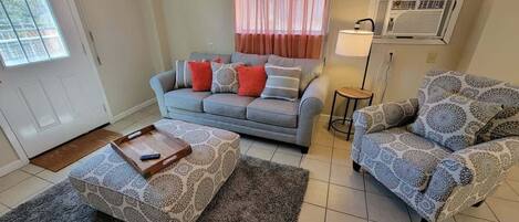 A living room with a grey sofa, patterned ottoman, and patterned armchair.