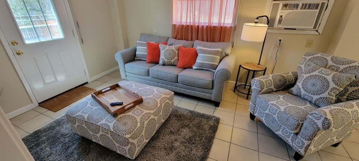 Un salon avec un canapé gris, un pouf à motifs et un fauteuil à motifs. On y trouve une petite table d’appoint avec une lampe.