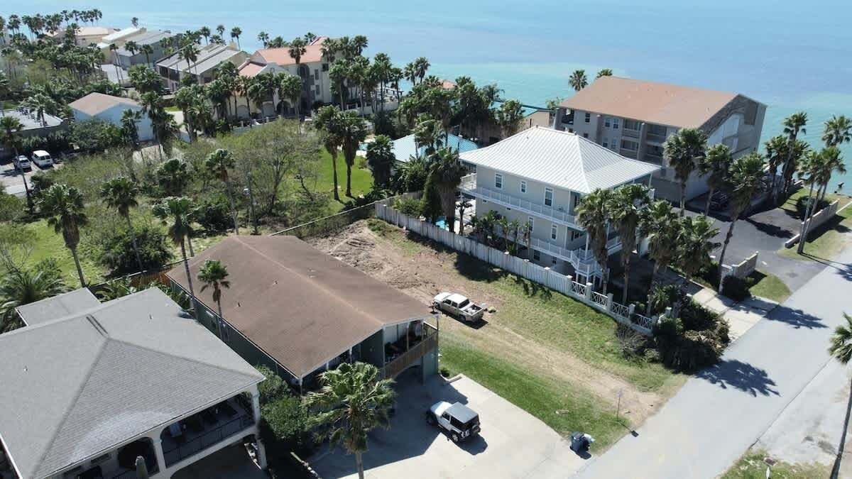 An aerial view of a coastal residential area with houses, a large building with a white roof, and palm trees.