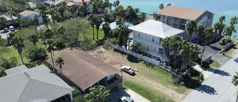 An aerial view of a coastal residential area with houses, a large building with a white roof, and palm trees.