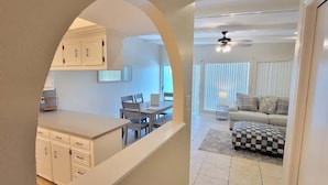 A kitchen with white cabinets and a dining area with a table and chairs. There is a checkered ottoman and a sofa in the living room.