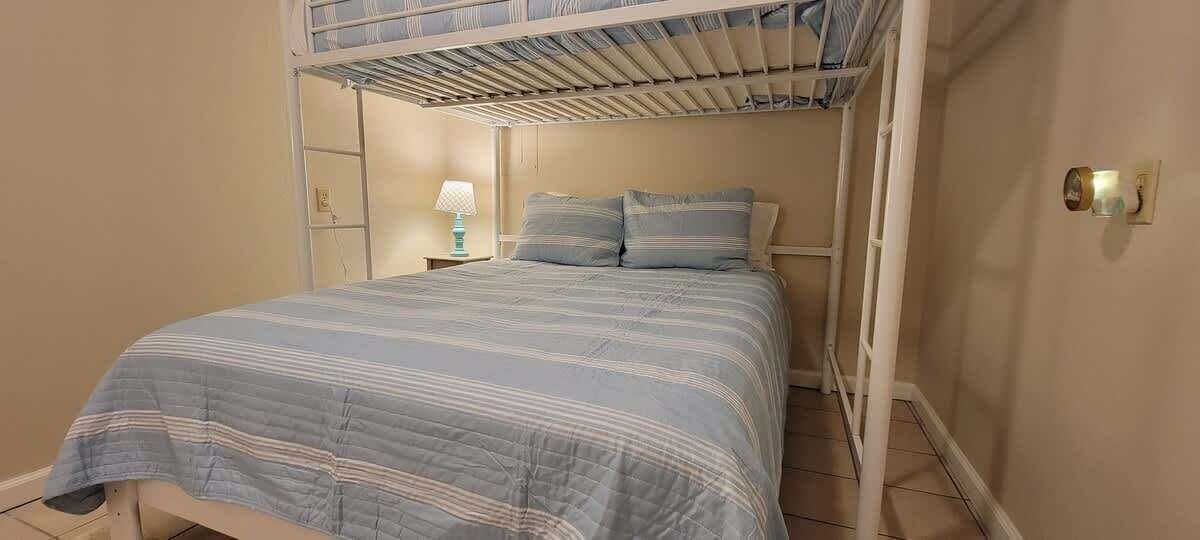 A single bed with a striped bedspread, a nightstand with a lamp, and a bunk bed above.