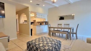 A modern living room with a sofa, ottoman, and dining area. A kitchen with white cabinets and a dining table is visible in the background.