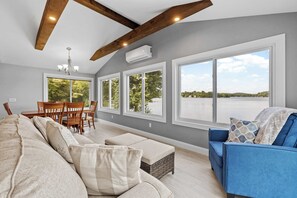 A modern living room with a large sofa, a blue armchair, a dining table with chairs, and large windows offering a view of a lake.