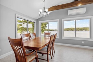 A dining room with a large wooden table, six chairs, a chandelier, and large windows with a view of trees and water.