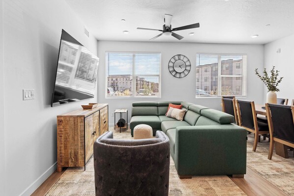 A modern living room with a sectional sofa, a flat-screen TV, a ceiling fan, and large windows with blinds.