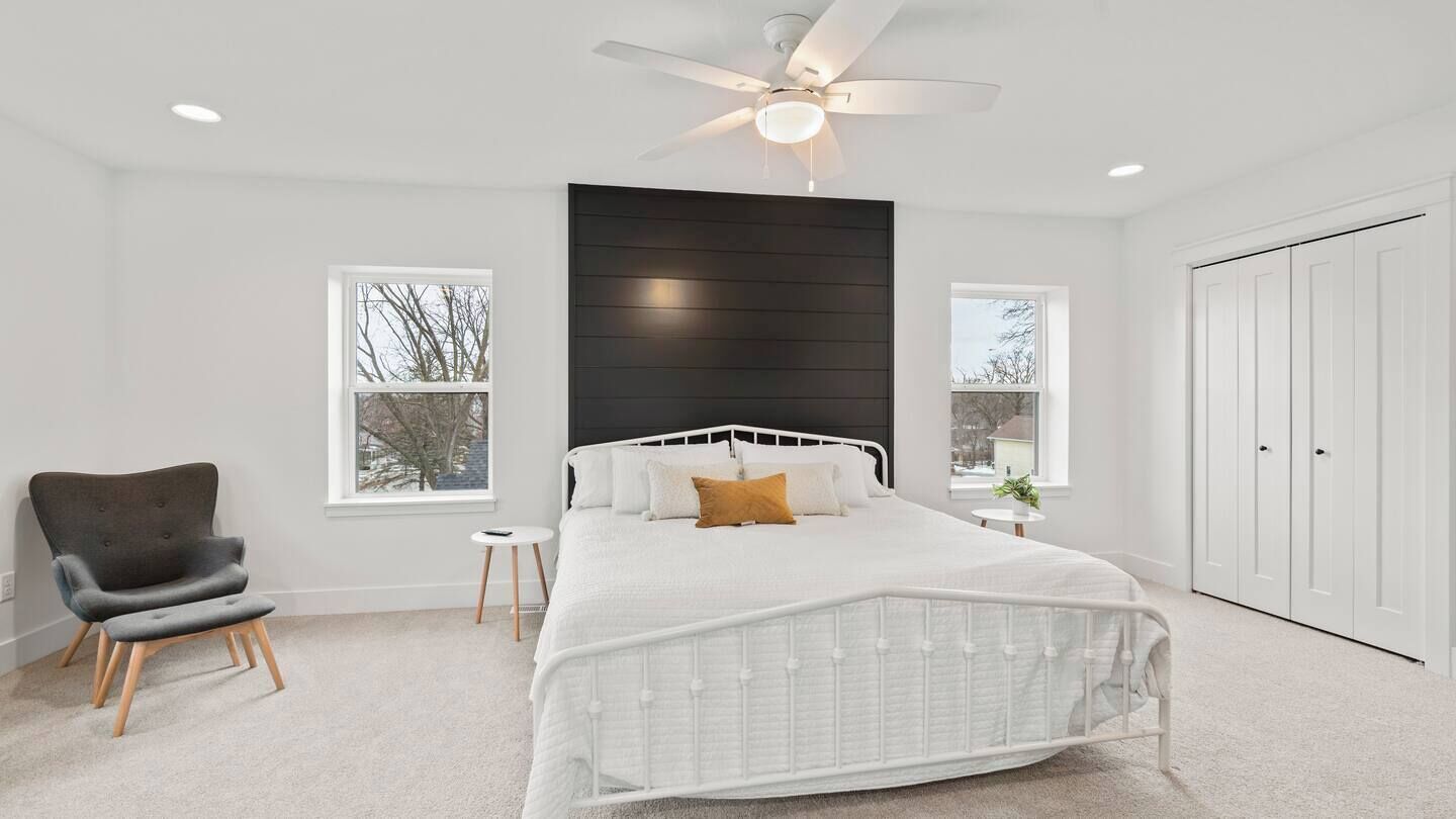 A bedroom with a white bed, a dark headboard, a ceiling fan, and a view of trees through the windows.