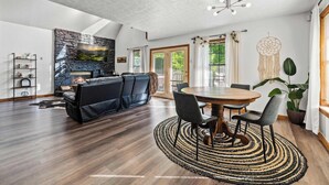 Un salon spacieux avec une cheminée en pierre, un canapé en cuir noir, une table à manger avec des chaises et une grande fenêtre avec des rideaux.
