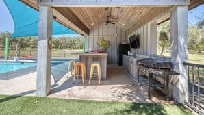 An outdoor patio with a barbecue grill, wooden stools, and a pool in the background.