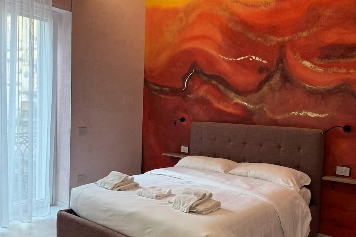 Un dormitorio moderno con una cama grande, un mural abstracto rojo en la pared y una ventana con cortinas translúcidas.